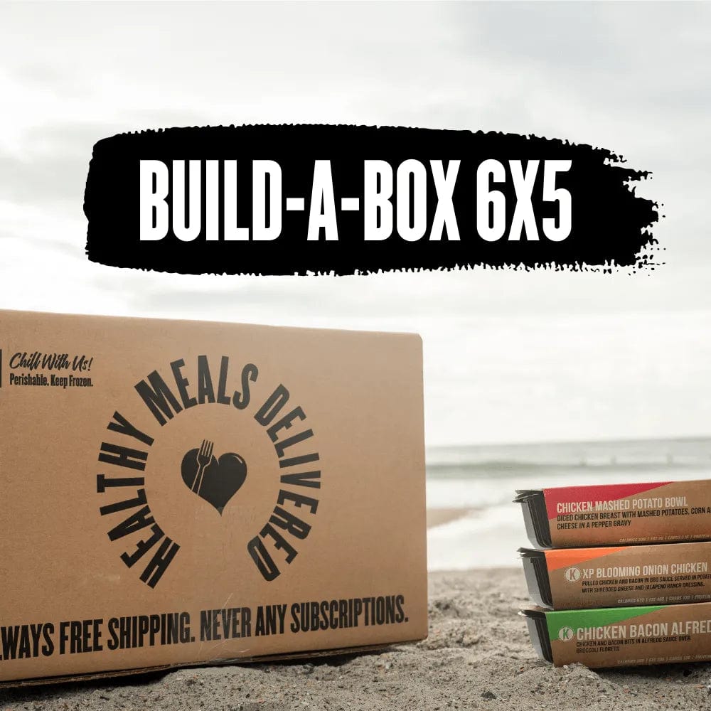 Clean Eatz Kitchen Build-A-Box 6x5 meal delivery package displayed on a sandy beach with the ocean in the background. The cardboard box is labeled 'Healthy Meals Delivered' with additional text: 'Always Free Shipping. Never Any Subscriptions.' Nearby, three meal trays are stacked, showcasing product labels for meals like Chicken Mashed Potato Bowl and Chicken Bacon Alfredo